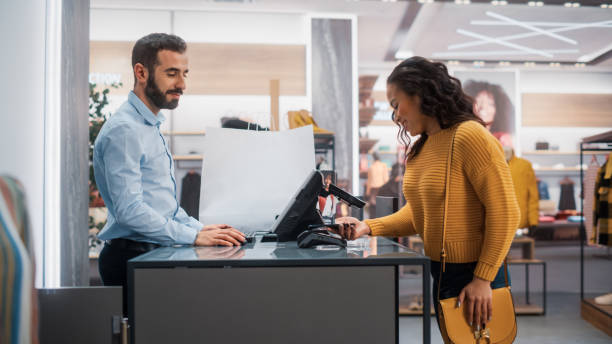 bekleidungsgeschäft: junge frau am schalter kauft kleidung von friendly retail sales assistant und bezahlt mit kontaktlosem nfc-smartphone-touching-terminal. trendiger modeladen mit designermarken. - shopping mall shopping sale retail stock-fotos und bilder