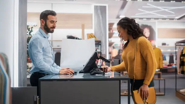 Photo of Clothing Store: Young Woman at Counter Buys Clothes from Friendly Retail Sales Assistant, Paying with Contactless NFC Smartphone Touching Terminal. Trendy Fashion Shop with Designer Brands.