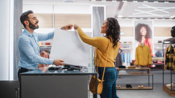 clothing store: young woman at counter buys clothes. friendly retail sales assistant gives her a shopping bag with new clothes. trendy fashion shop with designer brands. - butiksarbetare bildbanksfoton och bilder