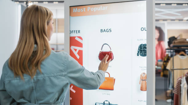 istock Beautiful Female Customer Using Floor-Standing LCD Touch Display while Shopping in Clothing Store. She is Choosing Stylish Bags, Picking Different Designs from Collection. People in Fashionable Shop. 1345105962