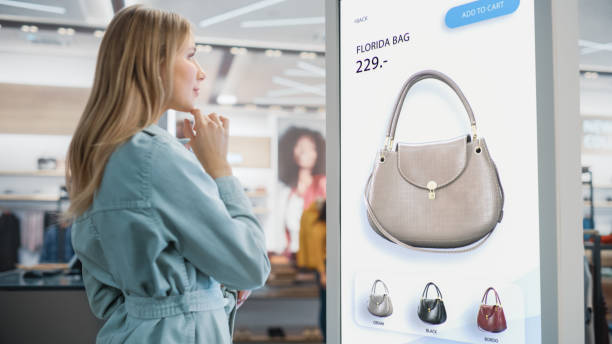 istock Beautiful Female Customer Using Floor-Standing LCD Touch Display while Shopping in Clothing Store. She is Choosing Stylish Bags, Picking Different Designs from Collection. People in Fashionable Shop. 1345105993