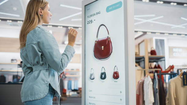 istock Beautiful Female Customer Using Floor-Standing LCD Touch Display while Shopping in Clothing Store. She is Choosing Stylish Bags, Picking Different Designs from Collection. People in Fashionable Shop. 1345105997