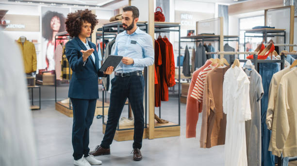 negozio di abbigliamento: una donna d'affari utilizza un tablet computer, parla con uno specialista di visual merchandising, collabora per creare una collezione elegante. small business fashion shop sales manager parla con il designer. - department store foto e immagini stock