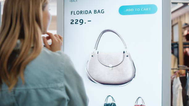istock Beautiful Female Customer Using Floor-Standing LCD Touch Display while Shopping in Clothing Store. She is Choosing Stylish Bags, Picking Different Designs from Collection. People in Fashionable Shop. 1345106007