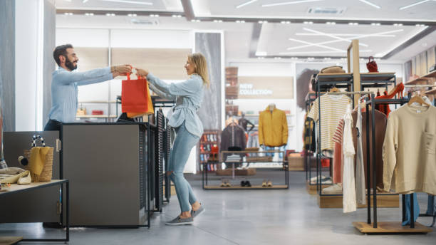 bekleidungsgeschäft: junge frau an der theke nimmt einkaufstaschen mit kleidung von friendly retail sales assistant, geht weg in happy mood, lächeln, spinnen und tanzen. modeshop mit designer-marken. - shopping mall store shopping retail stock-fotos und bilder