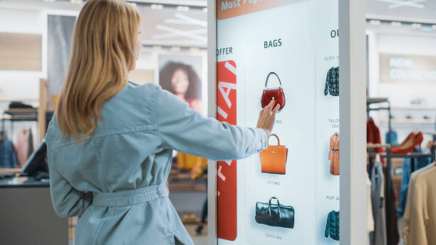 istock Beautiful Female Customer Using Floor-Standing LCD Touch Display while Shopping in Clothing Store. She is Choosing Stylish Bags, Picking Different Designs from Collection. People in Fashionable Shop. 1345106030