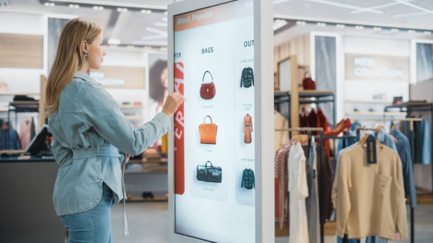 istock Beautiful Female Customer Using Floor-Standing LCD Touch Display while Shopping in Clothing Store. She is Choosing Stylish Bags, Picking Different Designs from Collection. People in Fashionable Shop. 1345106032