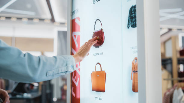 istock Beautiful Female Customer Using Floor-Standing LCD Touch Display while Shopping in Clothing Store. She is Choosing Stylish Bags, Picking Different Designs from Collection. People in Fashionable Shop. 1345106041