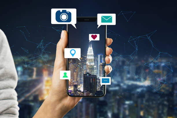 istock Close up of hand holding cellphone device with abstract polygonal mesh, camera, location pin, email and chat icons on night city background. Social media, communication, technology and internet concept. 1345107213