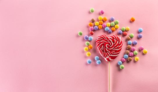 Heart-like striped red lollipop surrounded by sweet colorful candies laying on the pink background.