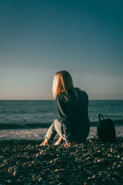 Photo of Young blonde woman is sitting on a pebble beach by the sea and looking at the sunset, a photo of her back. Concept of travel, enjoyment, loneliness. Vertical photo