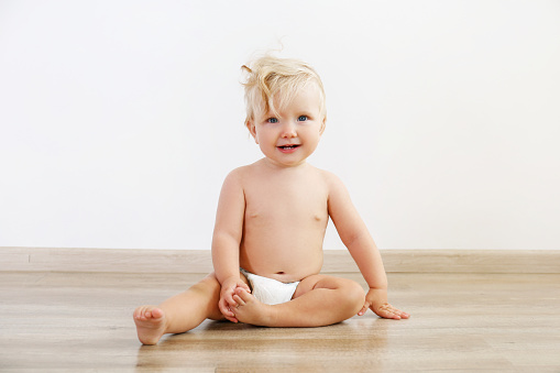 Portrait of one year old child sitting on the wooden floor over white wall background. Adorable blonde little girl in diaper. Close up, copy space for text.