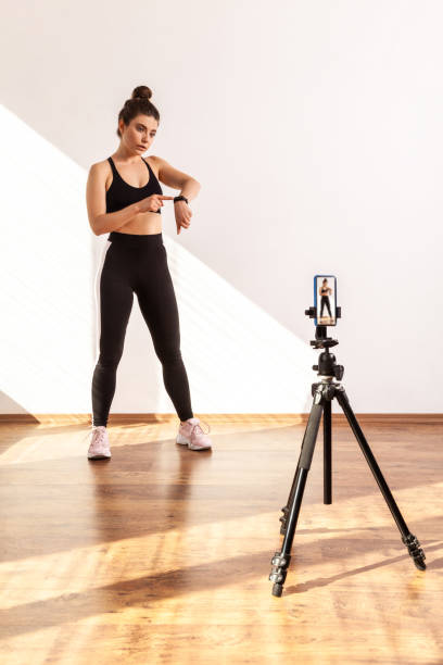 Serious athletic online trainer broadcasting livestream with workout, pointing at smartwatch.