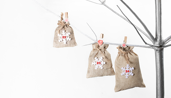 Bags made of matting for gifts with numbers 22, 23 and 24 on them hanging on clothespins on alternative Xmas Tree made of silver painted wood on white backdrop. 22 - 24 part of advent calendar.