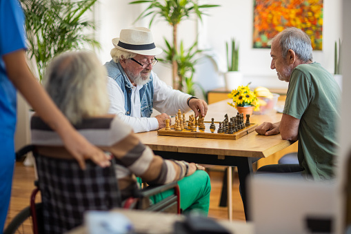 Senior men having fun playing chess at nursing home with nursing assistant beside them