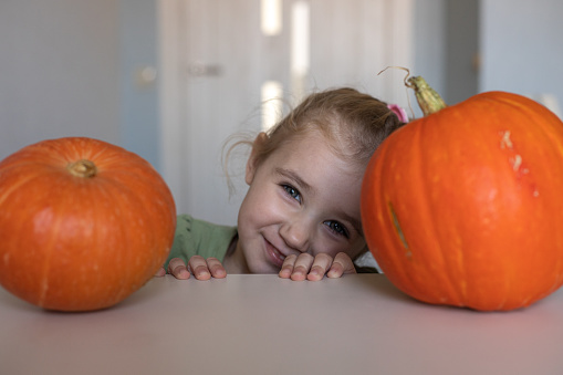 A beautiful blonde girl smiles at the camera at the table between two pumpkins in the kitchen