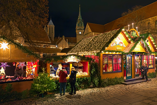 Braunschweig, Germany - December 7, 2018: Market stalls with Christmas decorations and sweets at Christmas market on Burgplatz square in dusk. Fragments of the Brunswick Lion Monument, Castle Dankwarderode, Town Hall and Brunswick Cathedral are visible in the background. Unknown people stand close to market stall with sweets and talk with unknown market vendor.