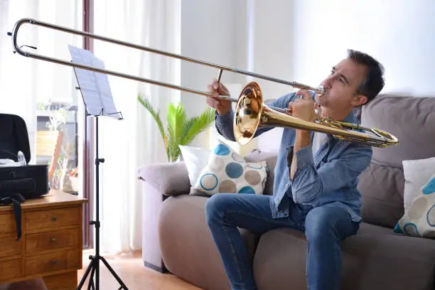 Novice man playing the trombone at home sitting on the sofa. Front view. Horizontal composition.