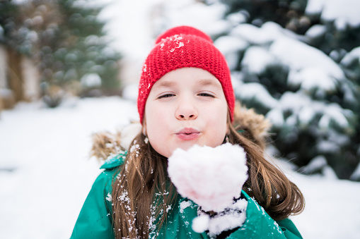 Wintertime. Cheerful caucasian girl in warm casual clothes blowing a handful of snow  on winter day. Kids having fun outdoor in cold weather.