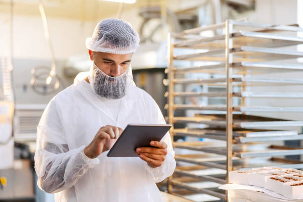 ein vorgesetzter in steriler uniform steht in einer lebensmittelfabrik und überprüft mit seinem tablet die produktivität. - hair net stock-fotos und bilder