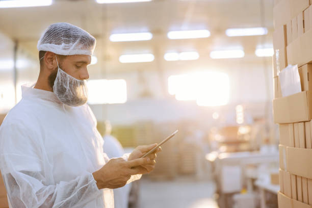 istock A supervisor with a hairnet and beard net in a sterile white uniform is standing next to a pile of boxes and checking them. He holds the tablet in hand and looking at it. 1345171794