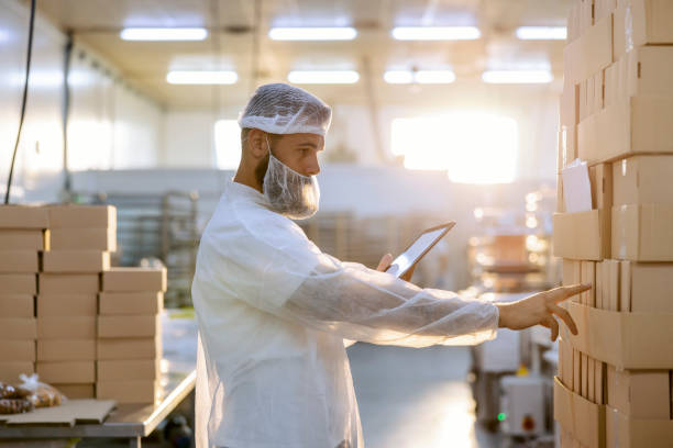 un joven inspector con uniforme blanco estéril está parado en una fábrica junto a cajas y contándolas. el número de cajas tiene que coincidir con los números de la tableta. -  producción y distribución de alimentos fotografías e imágenes de stock