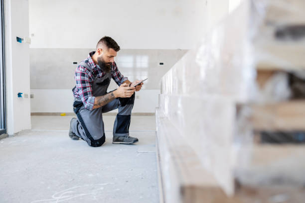 An innovative supervisor in overalls is kneeling inside the new building and checking on materials on his tablet. He is kneeling next to a pile of beams.