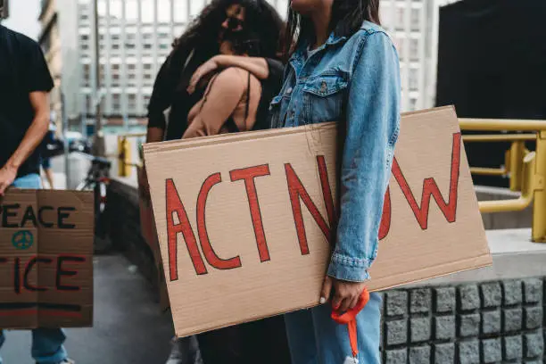 Detail of a young adult woman holding a cardboard sign with "act now" words on it. She's with other friends, they are going a demonstration against racism.