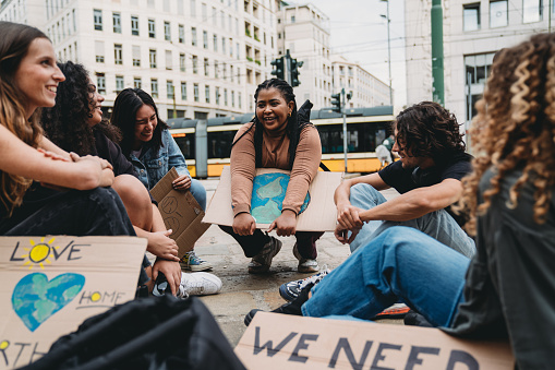 A group of young adult people are resting after a climate change demonstration in the city. They are sitting in circle with some cardboard signs against global warming.