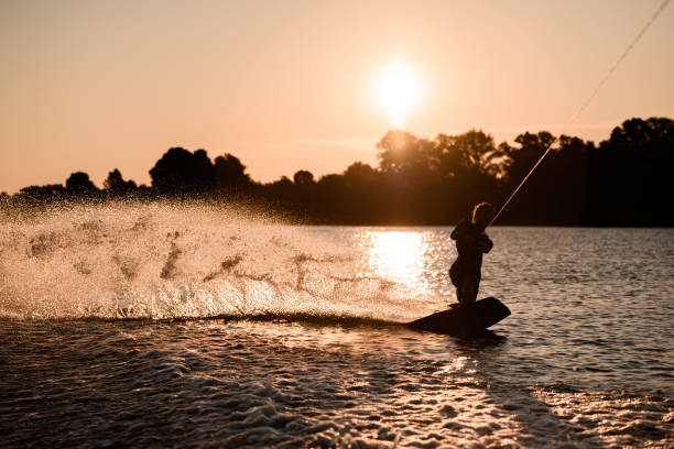 アクティブな男性ライダーの暗いシルエットの素晴らしいビューは、日没時に水面にウェイクボードに乗ってロープを保持しています。 - wakeboarding waterskiing water ski sunset ストックフォトと画像