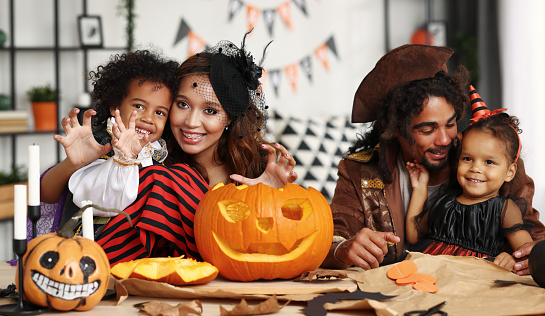 Portrait of happy african american family mother father and children smiling at camera while carving jack o lantern from pumpkin together, parents with kids preparing Halloween decorations at home