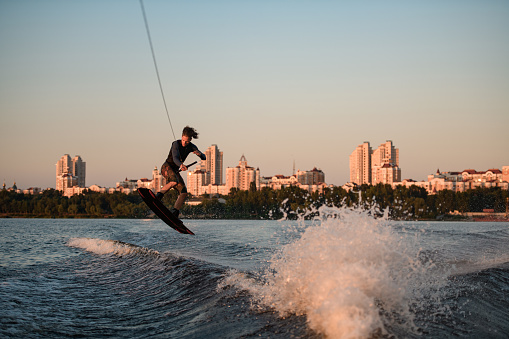 wonderful view of active male rider holds rope and masterfully jumping over splashing wave on wakeboard against the backdrop of city houses.