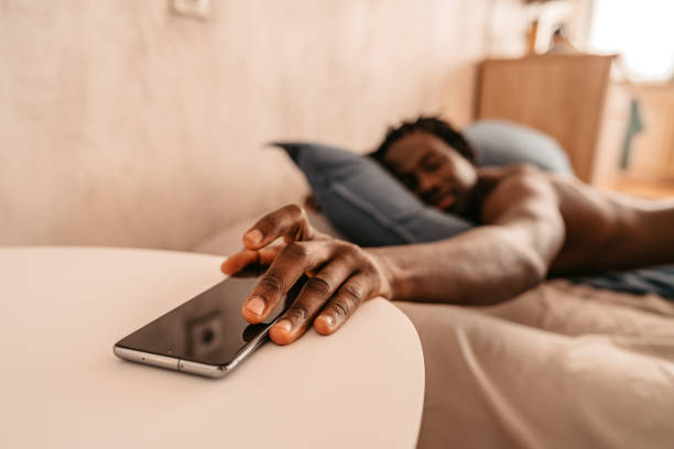 Man Snoozing Alarm On Mobile Phone Screen Tired and sleepy African-American man waking up in bed while turning off the alarm in his smartphone during the morning. oversleeping stock pictures, royalty-free photos & images