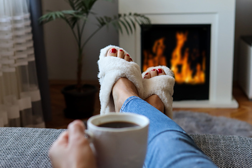 Female bare feet in white room slippers and warm legs next to the fireplace. A woman lies in bed and relaxing with a hot coffee drink