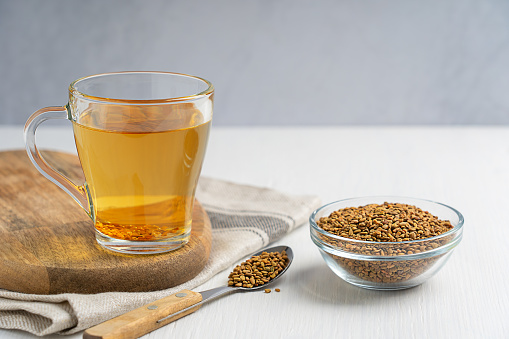Helba golden tea drink made of fenugreek annual plant served in glass cup with bowl of seeds and spoon on white wooden table with textile used in medicine of Egypt and as food ingredient. Horizontal
