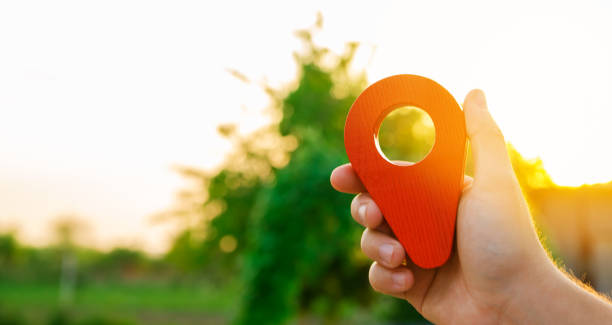 A hand is holding a red location marker in the sunset background. Navigation and exploration. The concept of tourism and travel. Destination. Holiday, vacation. Buying building land. Selective focus