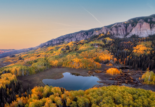 Drone photo of a small pond in the Colorado wilderness at sunrise surrounded by colorful yellow Aspens on a autumn morning.