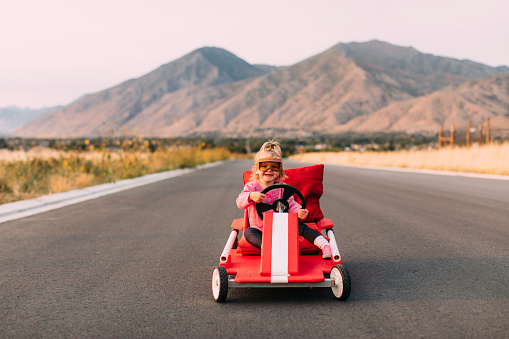 A young toddler girl sits in a homemade soap box racing car on a rural road in Utah. She is dreaming of hitting the roads and heading on a journey.