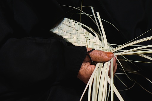 Hands of elderly arab woman artisan in traditional black dress close up weaving manually basket from dried organic palm leaves. Arabian traditional crafts.