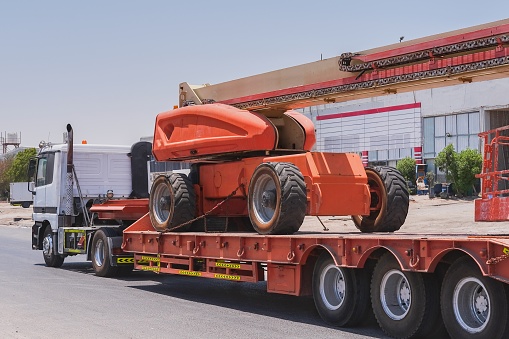 Transportation of old mobile tower crane with waste tyres to repair station on long cargo platform of truck trailer. Lorry delivery truck, cargo roadtrain for heavy machinery vehicles carriage.