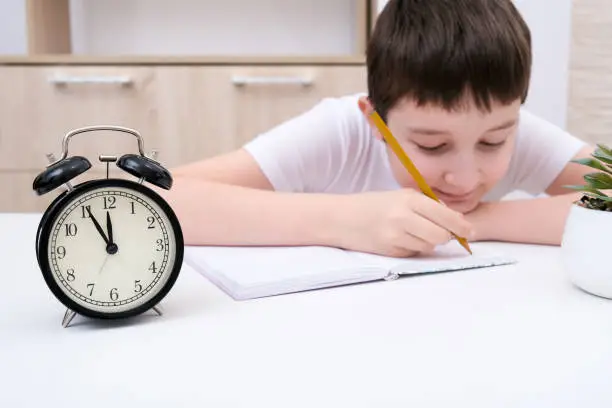 A concentrated boy writing in a textbook, exercisebook, a schoolboy doing his homework at home, an alarm clock in front of hin showing deadline.