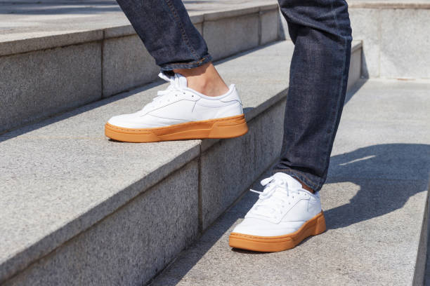 piernas masculinas en jeans y zapatillas blancas sobre fondo de escaleras de concreto. - zapato fotografías e imágenes de stock