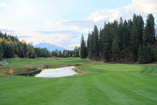 A beautiful view of of a fairway looking downhill towards a green with a lake beside it, on a beautiful golf course with autumn colours, in the rocky mountains near Canmore, Alberta, Canada