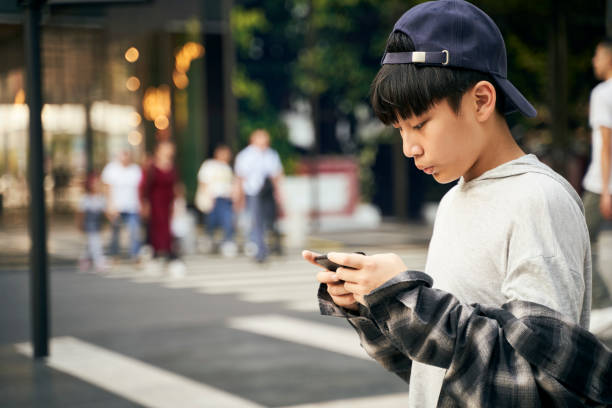 asian teenage boy playing with cellphone outdoors on street stock photo