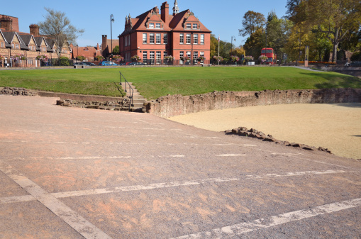 Markings showing the layout of the old Amphitheatre in Chester can be seen in the foreground with the old walls of the main pit and a grass bank seen in the background. In the distance are old Victorian era buildings.