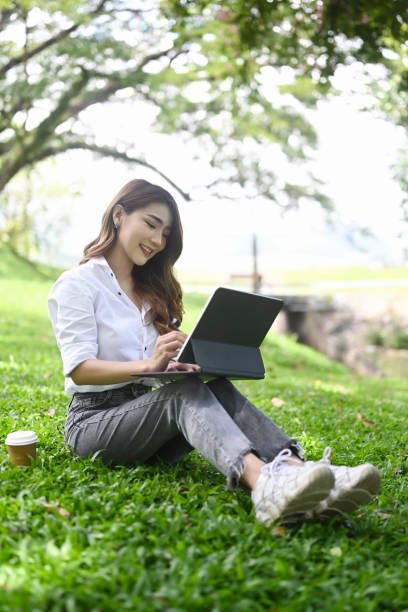istock Young female freelancer sitting on grass in public park and working with computer tablet. 1345256918