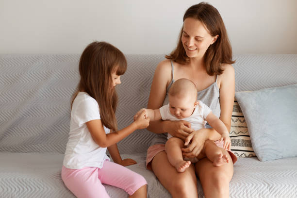 istock Happy mother with her cute daughters sitting on sofa at home, spending time together, elder sister holding infant baby hand, people expressing happiness. 1345258458