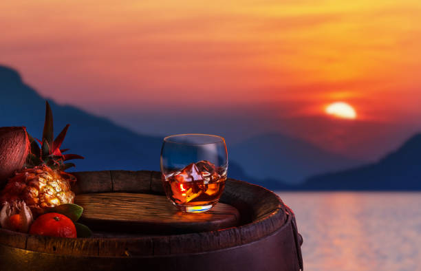 Glass of iced dark rum and tropical fruits over oak barrel. The red beautiful sunset sky at the sea shore. stock photo
