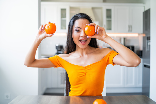 Beautiful and funny young brunette with orange shirt  holding two oranges in front of her face with toothy smile. Domestic background. Fun at home. Breakfast preparation