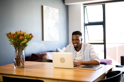 Wide shot man working at home on laptop smiling  to coworkers on video conference